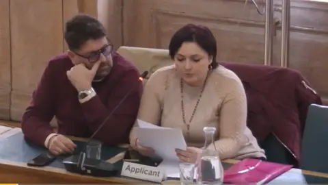 Kingston Council Alessandra and Vinvenzo Rea at the council meeting, with Alessandra holding up her notes and speaking into a microphone. She is wearing a cream white knitted top and her husband a ruby jumper with white shirt underneath.