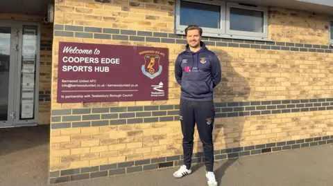 Barnwood AFC Chris Coward stands outside Coopers Edge Sports Hub, wearing a navy hoodie and black joggers. Winter sun is shining on him and he is smiling.