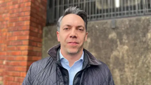 Dominic Muns looks at the camera standing in front of the grey concrete of the car park. He wears a blue padded coat and a blue shirt 