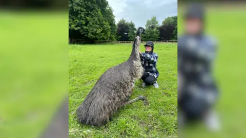 Rhi Evans Rhi Evans kneels in front of a fully-grown emu in a field with her hands on its neck. She is wearing a blue and white check shirt, dark leggings, white trainers and a black baseball cap. She is smiling at the bird.