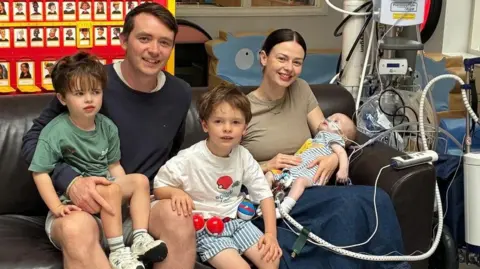 Family Photo Macs Green and family. His family are sat on a sofa. Macs is on his mother's lap. He is connected to hospital machinery