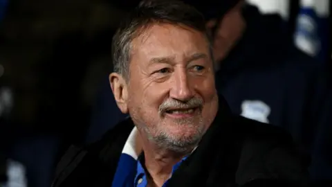 Steven Knight has short dark hair with flecks of grey, and a grey goatee. He is wearing a blue shirt, dark coat and a blue and white scarf. He is looking away from the camera to one side and smiling. 