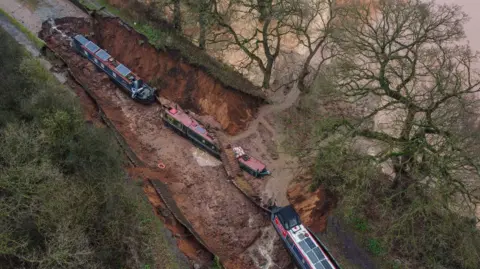 PA Media Three narrowboats seen from above, with two at the bottom of a muddy hole and a third on the edge, with water in an adjacent field