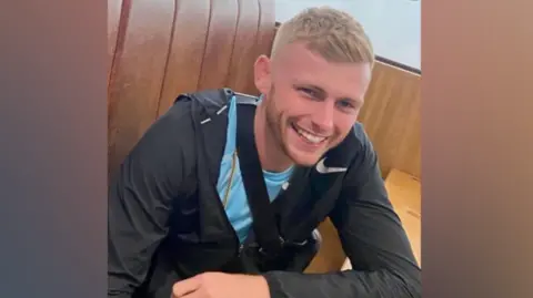 Family photo A man with blond hair smiles at the camera. He is wearing a light blue t-shirt and black jacket and has a bag strap on his shoulder.