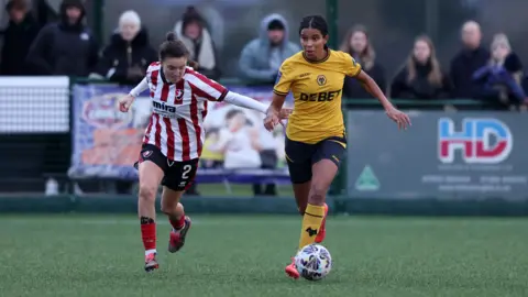 Getty Images Two women footballers running towards the ball with people in the background watching the match.