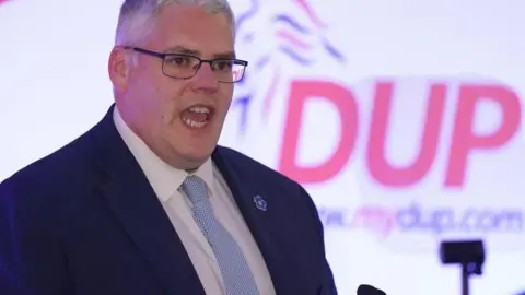 PA Media Gavin Robinson speaking at a DUP conference wearing a blue and white spotted tie, a white shirt and navy jacket. He is standing in front of a large, red letter DUP logo.