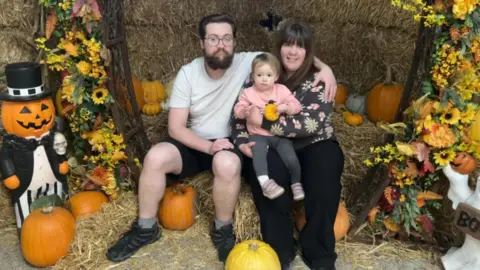 Lewis Relfe Lewis and Ameila Relfe sat down on some hay at a Halloween event. Ameila is holding their baby, Evelyn. There are pumpkins and sunflower decorations around them.