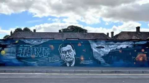 BBC A wall painted with a mostly dark blue mural, with roofs of houses behind the wall. The mural reads 'The Warminster Thing' and includes a flying saucer near a row of terraced houses. A portrait of journalist and ufologist Arthur Shuttlewood is included prominently.