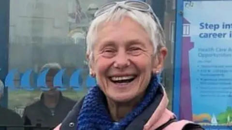 Dilys Gartside Dilys Gartside smiles broadly in a street outside an office. She has sunglasses pushed back over her short, white hair and wears a pink gilet and blue scarf.