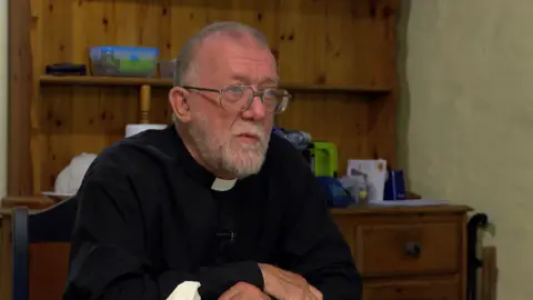 The image shows Reverend John Littlewood. He is wearing a black shirt with white clerical collar. He has short cropped hair and a beard and is wearing glasses. He is sitting in a room with a large wooden kitchen dresser behind him.