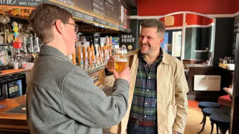 A man and a woman standing at a bar in a pub. They both have a beer and are making a cheers gesture with their glasses.