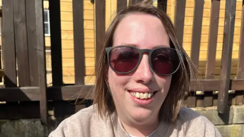 Emma Baugh/BBC Nicola is sitting in front of a brown wooden fence. She has short brown hair and is wearing a beige top and sunglasses.