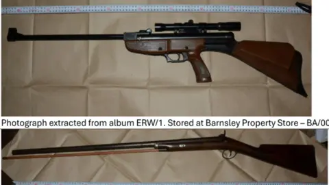 Crown Prosecution Service A composite of three separate photographs showing three long firearms laid horizontally on a sheet of brown paper. A white measuring scale runs parallel to each firearm, indicating that the weapons have been photographed for documentation or evidential purposes. In the top photograph, a rifle with a dark wooden stock and a black metal barrel is displayed. The firearm has a mounted telescopic sight positioned above the receiver. The barrel is relatively long and extends to the left side of the image, while the wooden butt stock is on the right. A pistol-style grip is incorporated into the stock design. The firearm is placed on a creased brown paper surface, and a measuring scale is visible along the upper edge of the photograph. In the middle photograph, a traditional long gun with a dark wooden stock and fore-end is shown. The firearm has a single long barrel extending almost the full width of the image. The stock and trigger area are located on the right side, while the muzzle points toward the left. A measuring scale runs along the bottom of the photograph. The firearm has a classic sporting-gun appearance with a simple wooden finish and metal barrel.