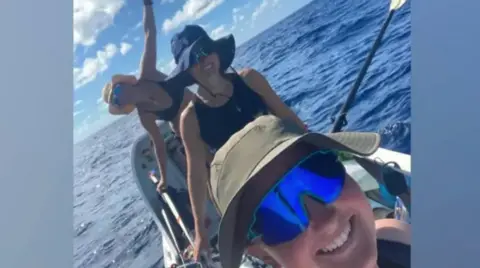 Row with the Flow/World's Toughest Row The picture shows three smiling people on a small ocean rowing boat in the middle of a vast stretch of deep blue water. One person is leaning out to the side of the boat with an arm raised, while another is seated and holding an oar. All are wearing sun hats. The sky is bright with scattered clouds, and the sea looks calm but expansive, giving a sense of isolation and adventure far from land.

