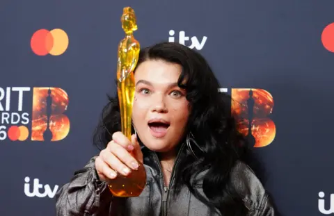 PA Media Lola Young holding up the gold Bris statuette in front of her with a shocked expression on her face