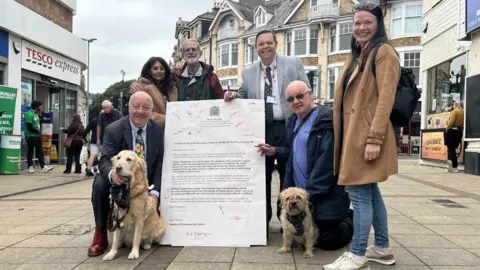 Guy Henderson/LDRS Several people gather on a street outside some shops including a Tesco Express. The group is gathered around an enlarged letter calling for urgent investment at Torbay Hospital. L-R: Steve Darling and his guide dog Jennie, an unknown woman in a brown coat, councillor Nigel Penny, councillor Swithin Long, unknown kneeling man with dog and councillor Cat Johns.