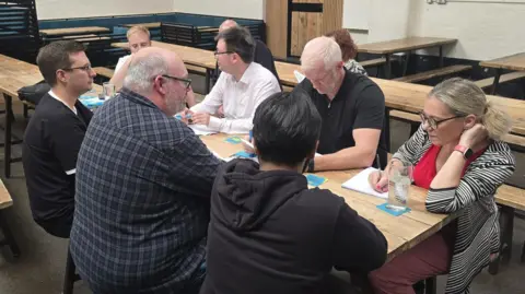 A group of people sit around a bench in a brewery, with several writing in notepads.