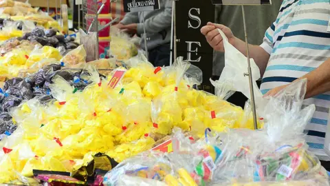 Man serving in front of a stall filled with yellowman in bags, alongside other sweets