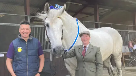 Luke Deal/BBC Luke Deal smiles at the camera wearing a navy gilet jacket and purple T-shirt. Next to him is Boysie the horse who is white and has blue decorations in his mane as well as a blue sash around his neck. Lynsey Atkin holds Boysie by the reins at his mouth. She wears a riding jacket, red tie and flat cap.