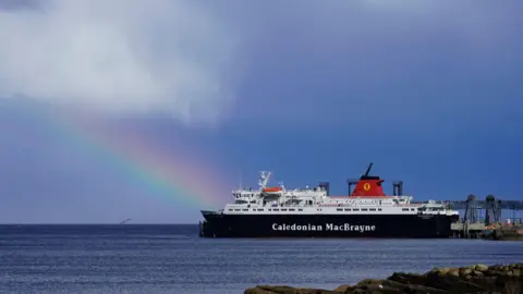 Martin Dean White and black CalMac ferry with a red funnel sailing. There is a rainbow behind the ferry and some clouds in the sky.