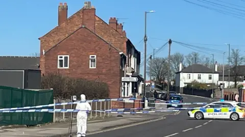 A street is taped off with a police car parked in the middle of the road. A scenes of crime officer in a white suit stands at the scene.
