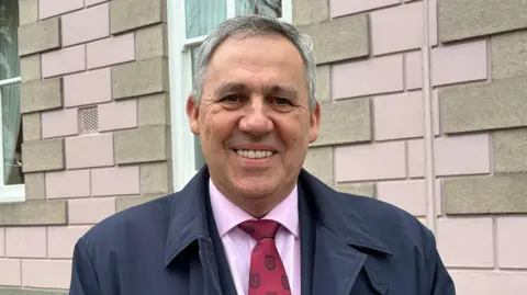 Constable Andy Jehan, a man with short grey hair. He is smiling directly at the camera, while standing in front of Jersey's States Chamber. He is wearing a pink shirt, maroon tie and a dark coat.