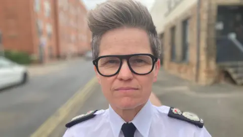 Rachel Swann wearing a white police uniform, black glasses and a large fringe hairstyle
