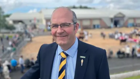 NFU President Tom Bradshaw at Devon County Show. He is wearing glasses, a blue suit and has a dapper blue and yellow striped tie on. 