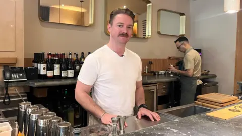 Chrissie Reidy/BBC Jake behind the bar with bottles of wine and a member of staff working beside him.