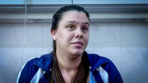 Mauren Echevarría Peña is sitting on a hospital bed. She is wearing a blue and white tracksuit. Her dark hair is pulled back in a ponytail, some of the hair falls over her shoulders.