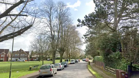 A road which has a field of grass to one side and a hedgerow on the other. There are parked cars on the common side. 