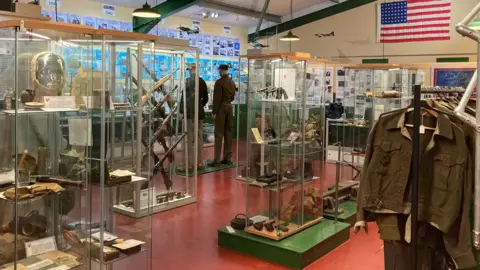 Katy Prickett/BBC The interior of the British Resistance Museum at Parham Airfield. Glass cases show an array of artefacts including decommissioned pistols, rifles, ammunition and other decommissioned armaments. A row of World War Two uniforms are hanging on a rail on the right. The walls are covered in information boards, telling the story of the museum. On the right is a US flag - Parham was home to the US Air Force at one point during World War Two.
