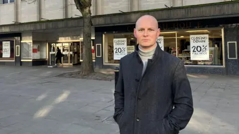 Max Wilkinson in front of a shop. He is wearing a grey coat.