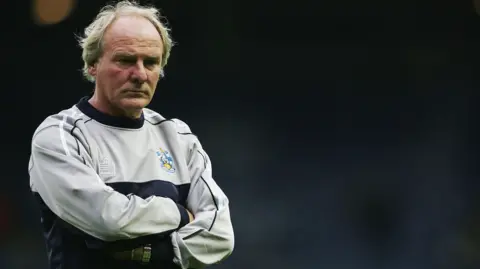 Getty Images Terry Yorath, pictured wearing a Huddersfield Town-branded tracksuit top and standing with his arms folded.