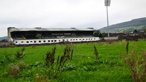 Overgrown grass in front of a stadium. Hills are in the background. 
