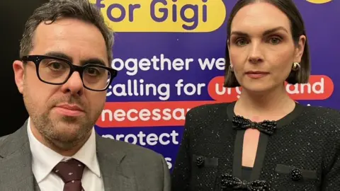 John and Katie Meehan. John has short black hair and is wear a grey suit with a burgundy tie while his wife has dark brown bobbed hair is wearing a black glitter suit jacket with black bows on it. They are standing in front of a purple yellow and red poster which says 'For Gigi'. They look serious.