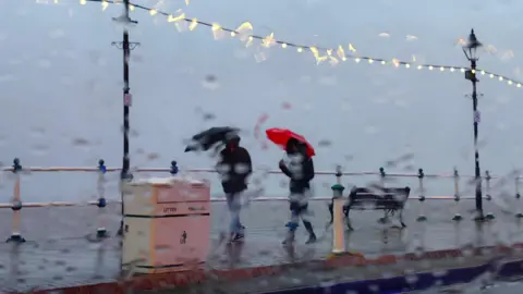 BBC Weather Watchers A photo taken through a window pane blurred with rain, showing two people walking under umbrellas