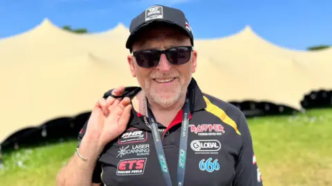 Ian Brindle is wearing a black racing polo shirt, a black cap and sunglasses. He grey stubble and is smiling, holding a jacket over one shoulder and stood on grass in front of a large teepee tent on a sunny day.