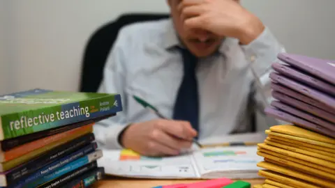 Teenage boy in shirt and school tie writing in a workbook at a desk with pens and other work books piled on it