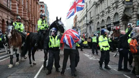 EPA/Shutterstock Police officers on horseback and with helmets. A man with a union jack flag wrapped around him is near to the officers. A crowd of people holding signs can be seen in the background.