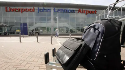 An outside shot of Liverpool John Lennon Airport with a suitcase in the right hand corner of the shot 