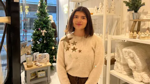 A woman wearing a grey jumper with star symbols on the shoulder standing inside a shop. The hop has white panelled walls and white display shelves. Homeware items including ornaments and blankets are on the shelves. A Christmas tree is on display in the shop window behind the woman. 