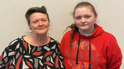 Suzanne Buckner Suzanne Buckner and her daughter face the camera. The mother has a patterned black, grey, red and white blouse and brown hair tied back in a wavy style. Her daughter has braided, brown hair and wears a red sweatshirt bearing the word "nemesis".
