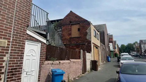 Simon Thake A quiet street with cars parked up down one side and a row of old houses with some patchy brickwork