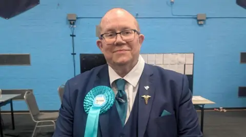 A man in a navy blue suit and tie is smiling at the camera, he is wearing glasses and a bright blue rosette
