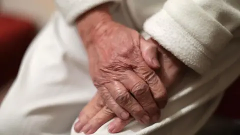 Anonymous close-up image of an elderly person with their hands crossed on their lap. They have white skin and they are wearing a white fleece top and pale trousers. 