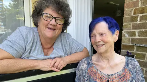 Marie in a grey t-shirt smiles over her first floor balcony alongside Thelma, with bright blue hair on her right.