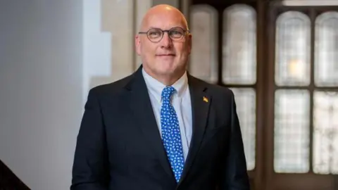 Hampshire County Council A press shot of Nick Adams-King posing on a wide stairway inside a building. He is wearing round glasses, a dark suit and a blue tie.