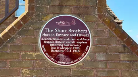 Steve Ladner/BBC A sign dedicated to the 'Short Brothers' Horace, Eustace and Oswald. The sign states the brothers were "aviation prioneers and their workforce founded Britain's aircraft seaplane and flying boat industry". The sign is maroon and was erected in 2008.
