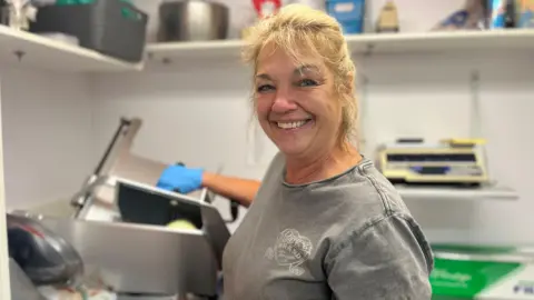 Kim is smiling into the camera. She's wearing blue gloves and a grey t-shirt and has blonde hair. Her right hand is reaching towards a slicing machine in the kitchen of a diner. the machine is silver and metallic.